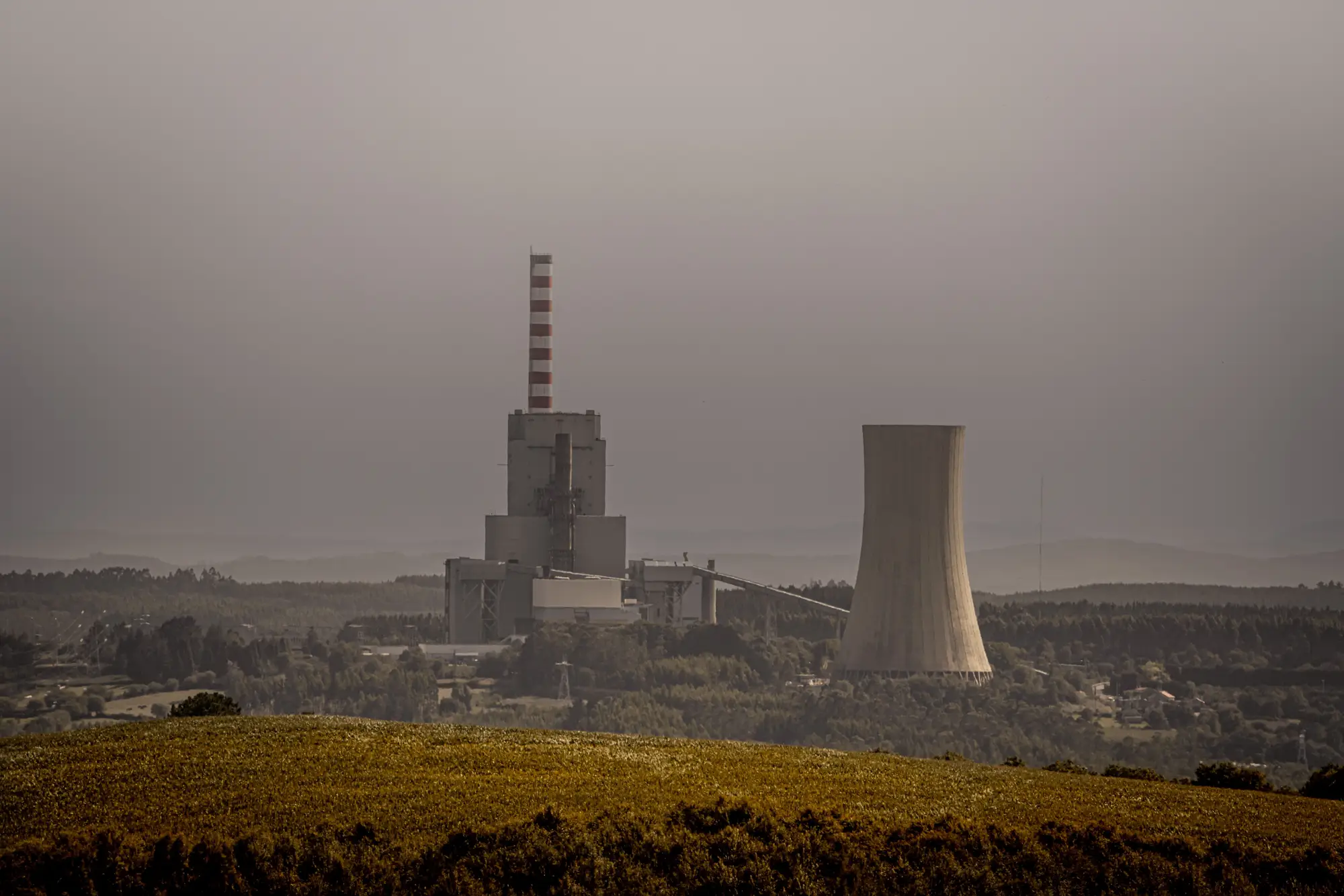 A coal-fired power station with its stack and cooling tower, seen from a hillside on a hazy day